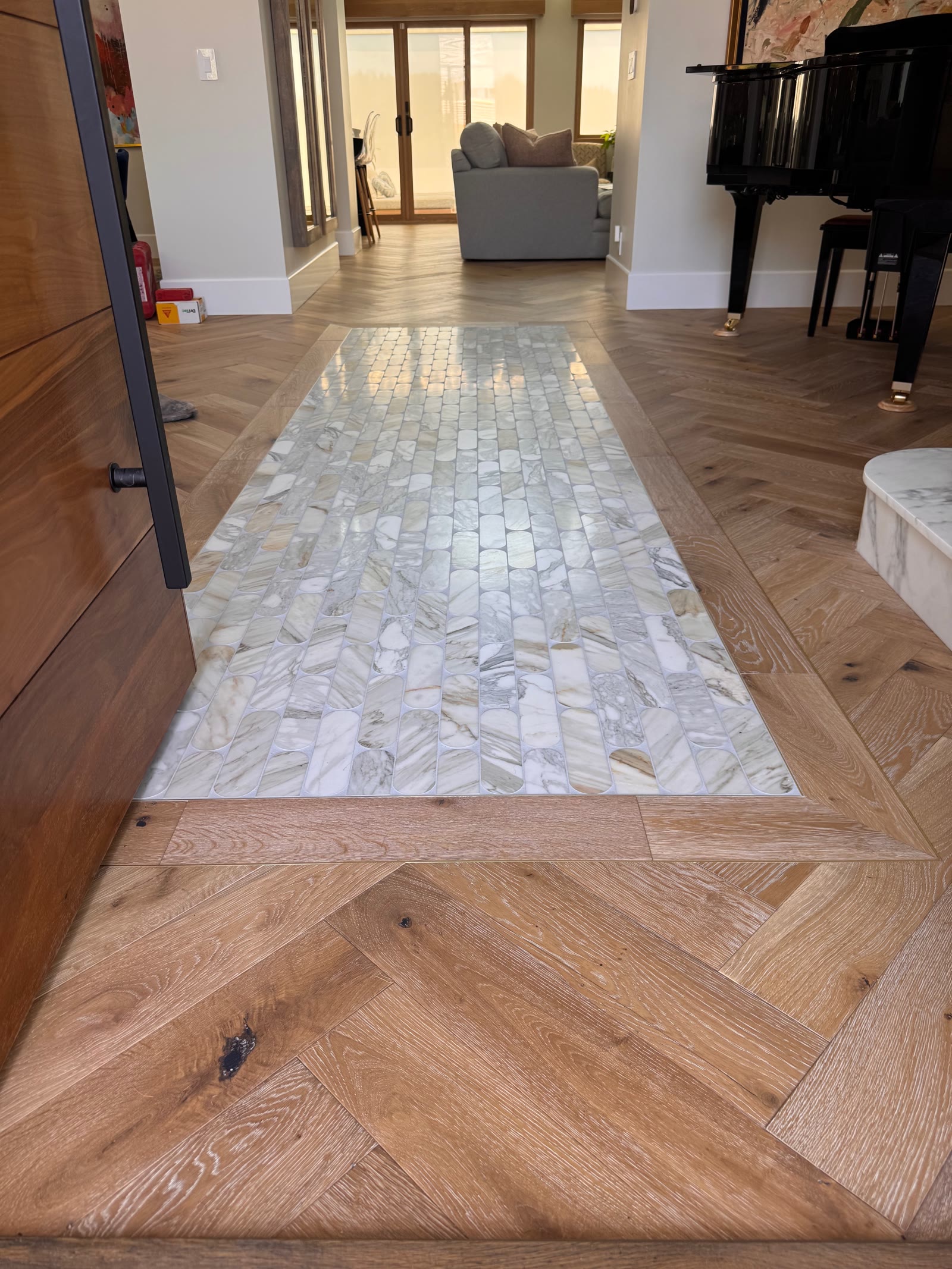 Herringbone white oak floor with a marble mosaic inlay runner set flush into it, beside a grand piano